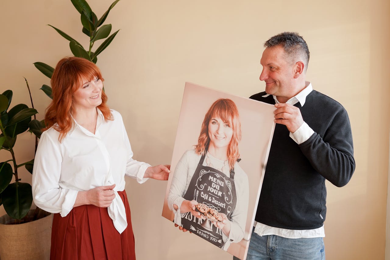 A happy couple smiles at a large personalized portrait they hold indoors.